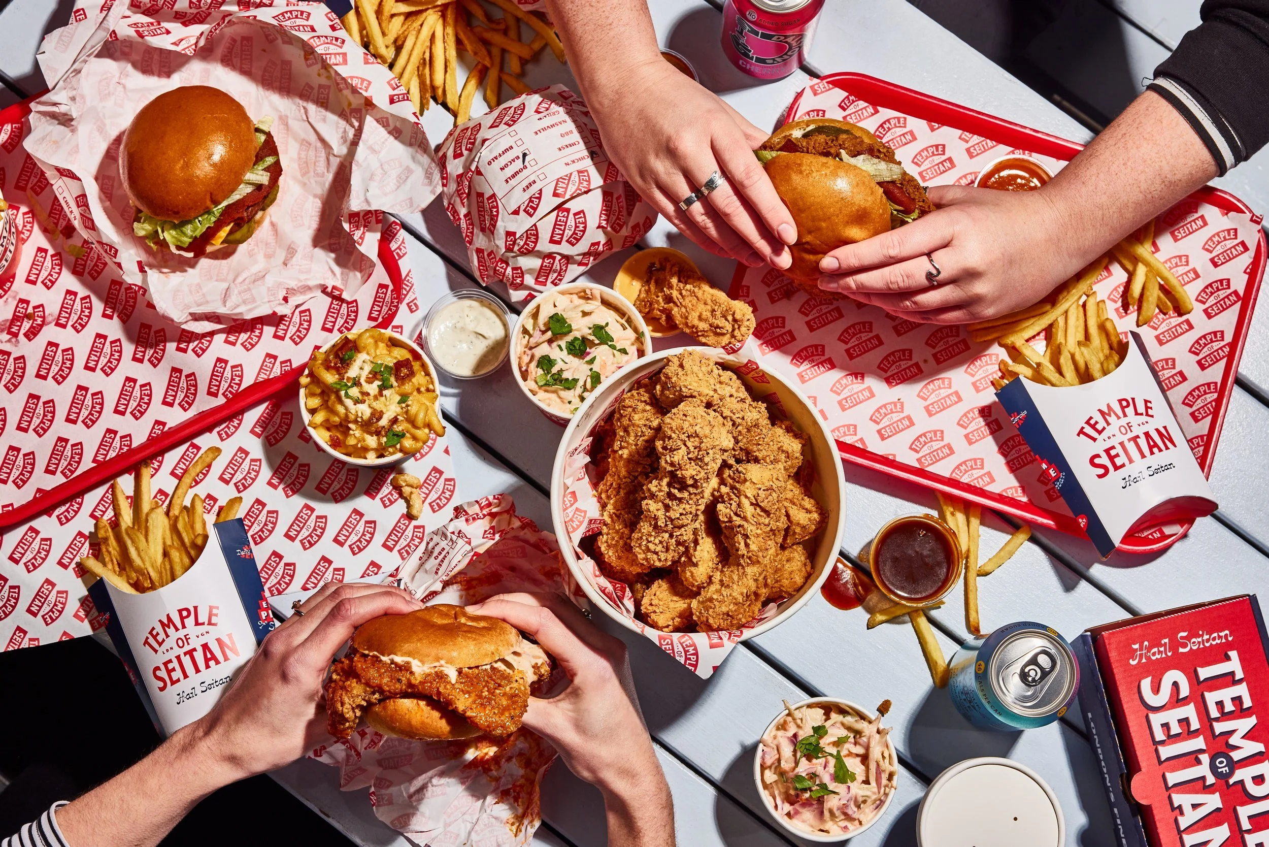 A table filled with fast food including fried chicken sandwiches, French fries, cole slaw, macaroni and cheese, fried chicken pieces, and several beverages. The table has red and white branded paper and bowls from Temple of Seitan.