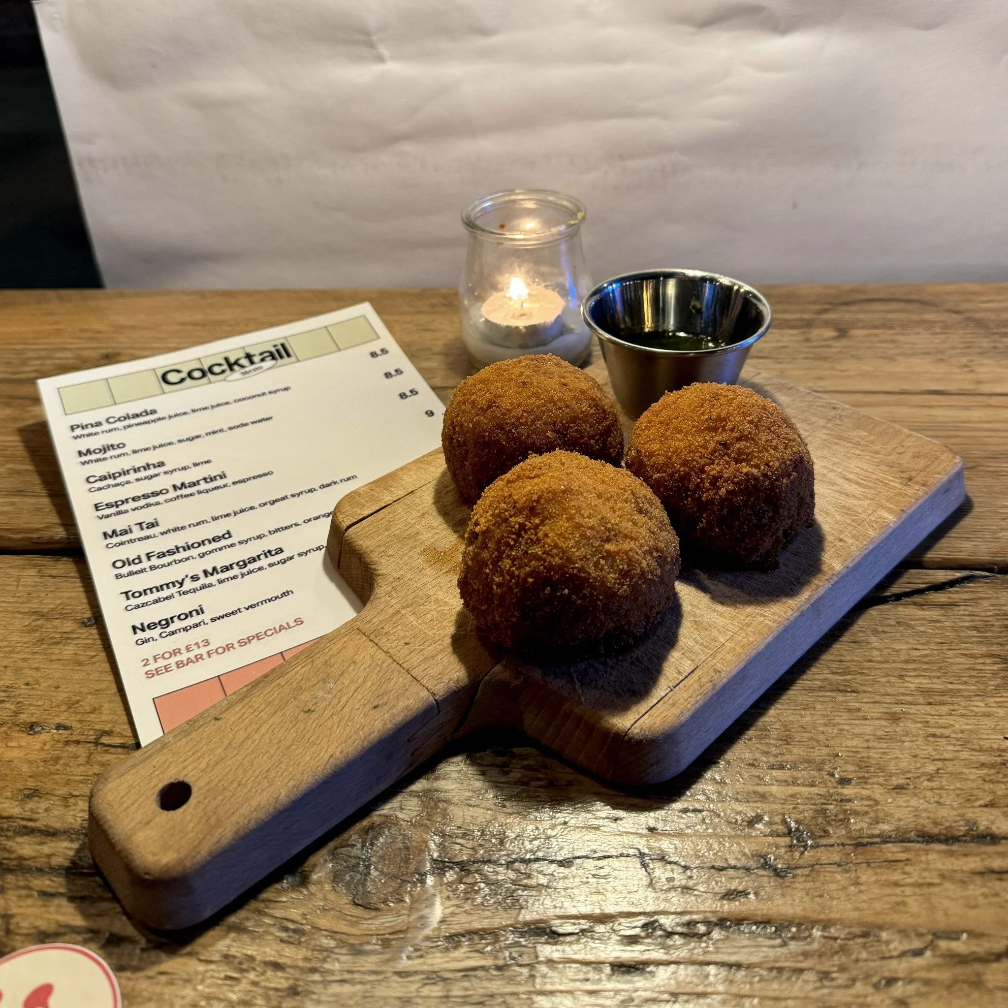 Fried food balls on a wooden serving board with a dipping sauce and a candle in a glass holder on a wooden table.