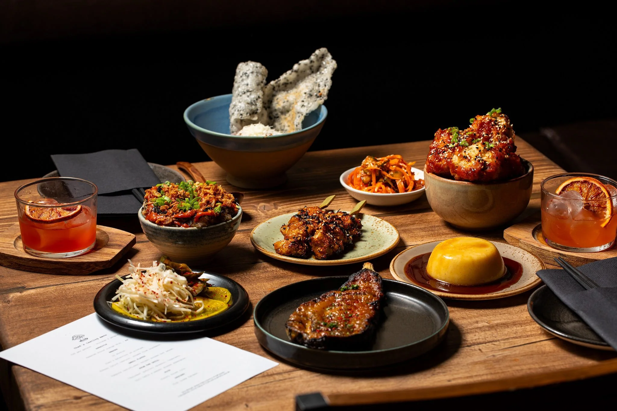 A spread of various Asian dishes on a wooden table, including bowls of rice, spicy chicken, grilled fish, vegetables, and drinks, with black chopsticks and a menu in the corner.