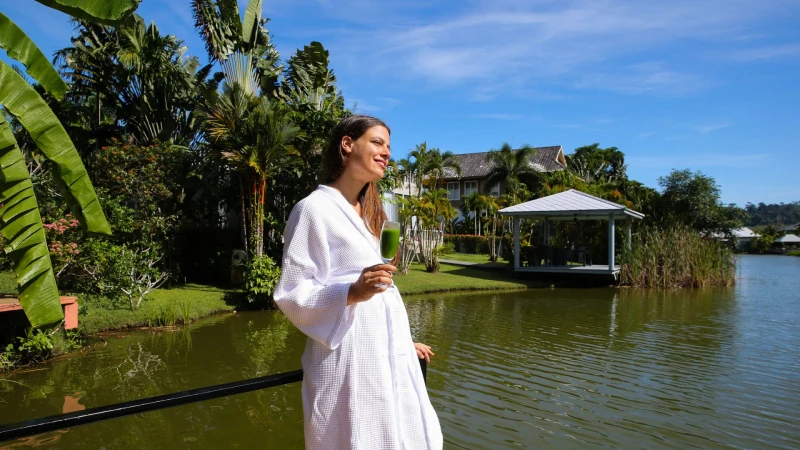 Woman in a white robe holding a green drink, standing by a lake with lush greenery and a gazebo in the background on a sunny day.