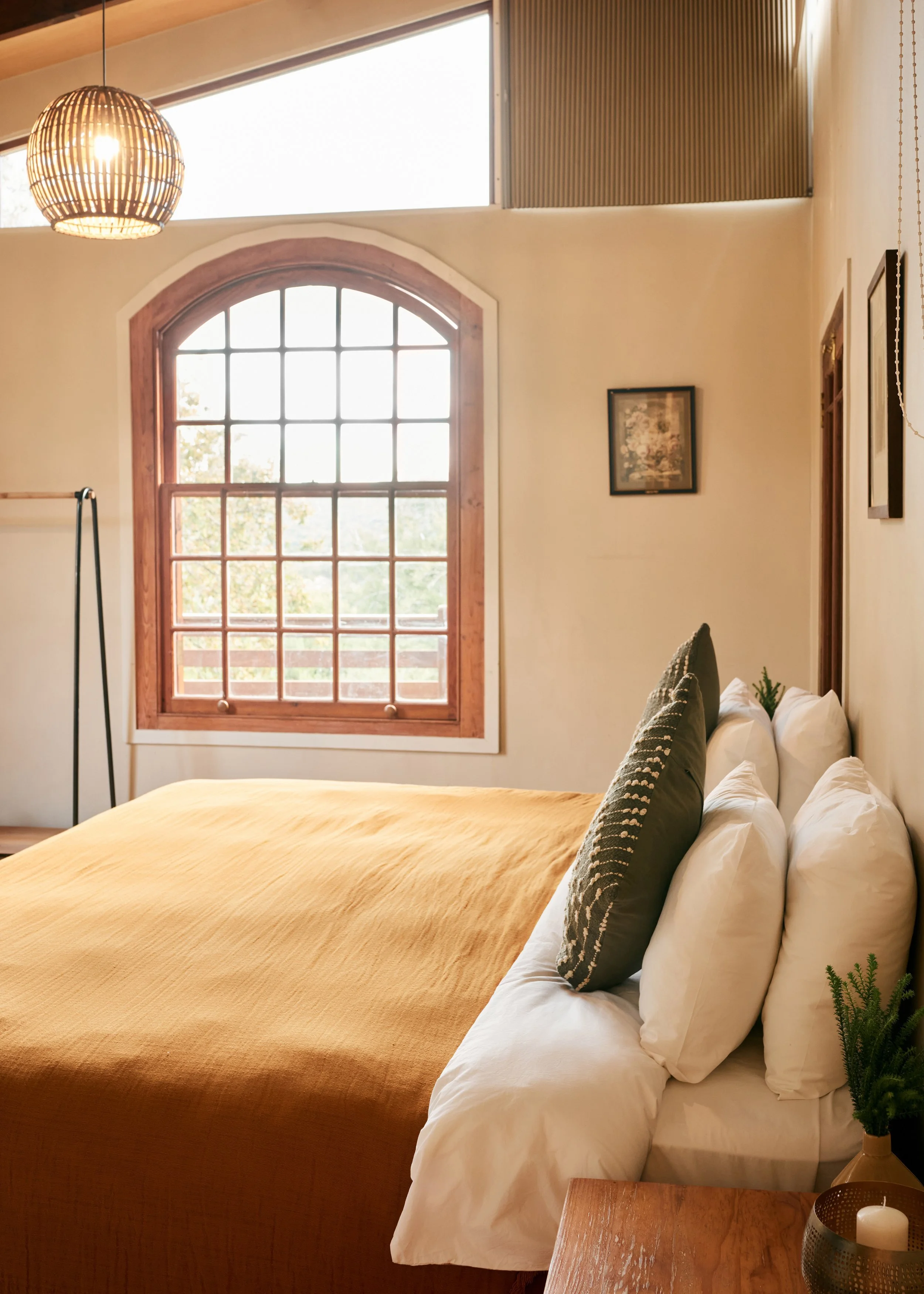 Modern bedroom with a large window, wooden accents, a bed with white sheets and yellow duvet, and gray pillows. There are framed pictures on the wall and a bedside table with a candle, a small plant, and decorative items.