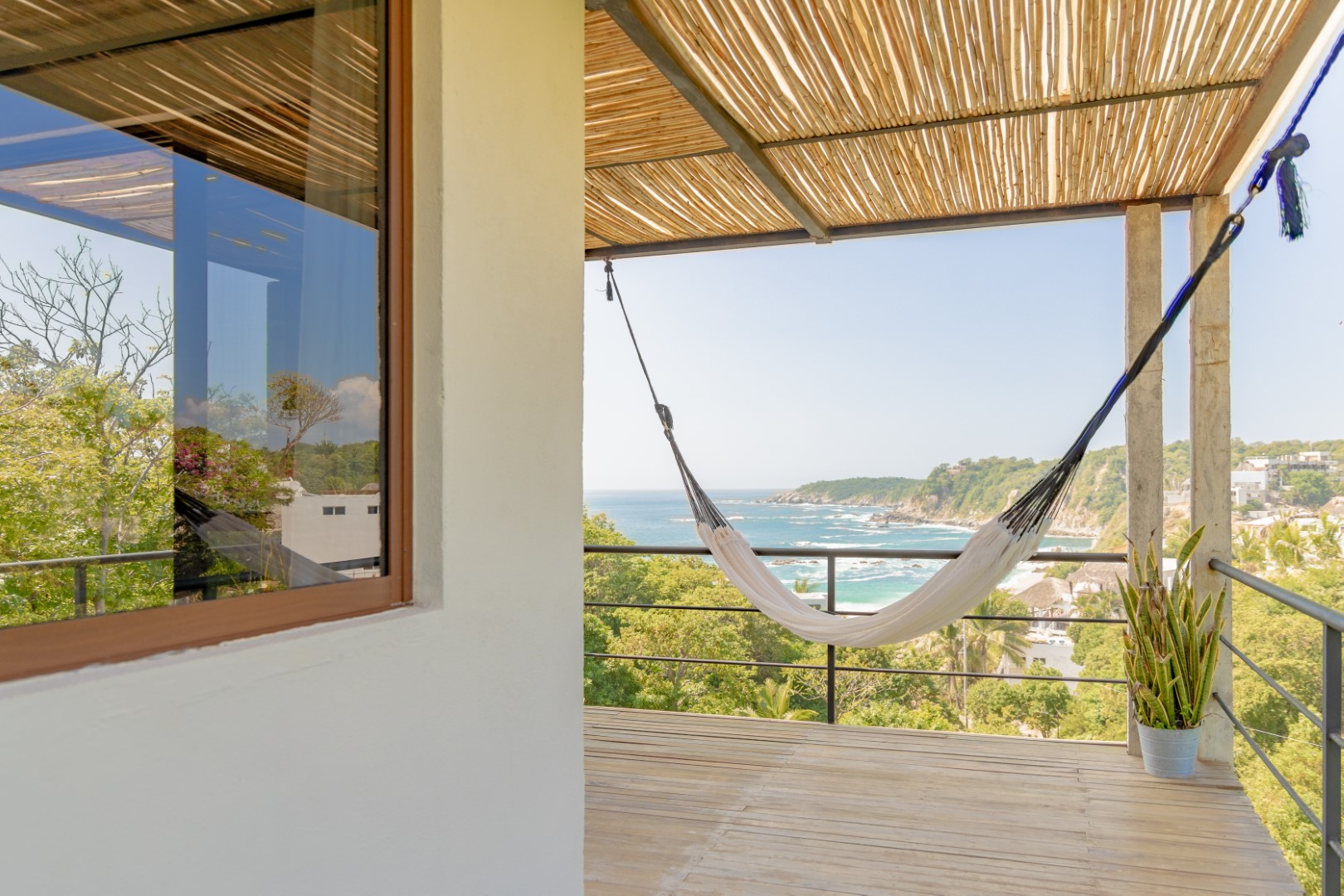 Balcony with a hammock, potted plant, view of ocean, cliffs, trees, and houses in a coastal area, under a straw roof.