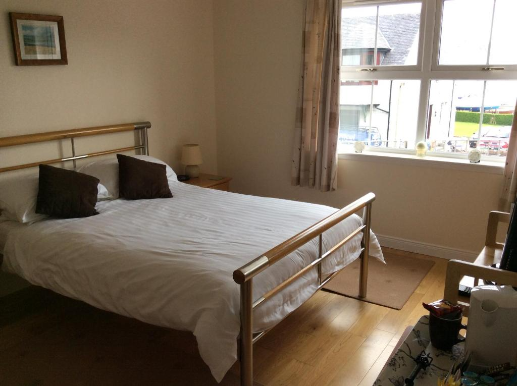 A bedroom with a metal bed frame, white bedding, and two dark brown pillows. There is a small bedside table with a lamp, a window with curtains, and part of a sitting area with a chair and a table with objects on it.
