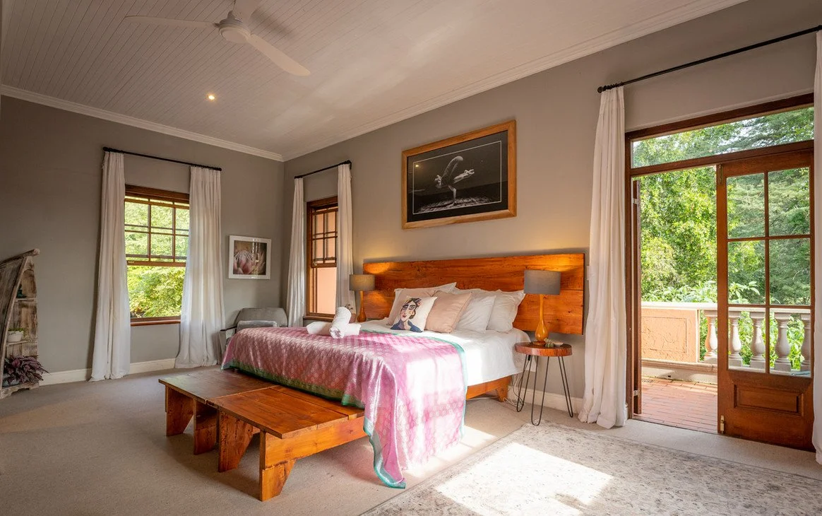 A bedroom with a wooden bed frame, white bedding, and pink throw blanket. Two nightstands with lamps, framed artwork on the walls, and windows with white curtains. An open door leading to a balcony with a view of green trees.