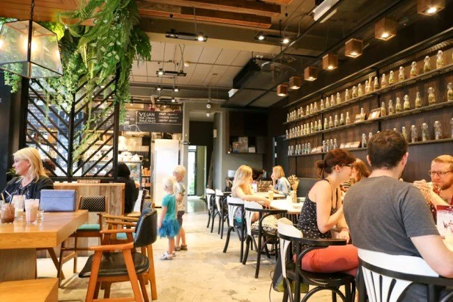 Inside a vegan restaurant with customers dining at tables, shelves with jars on the wall, and a small child walking through the space.