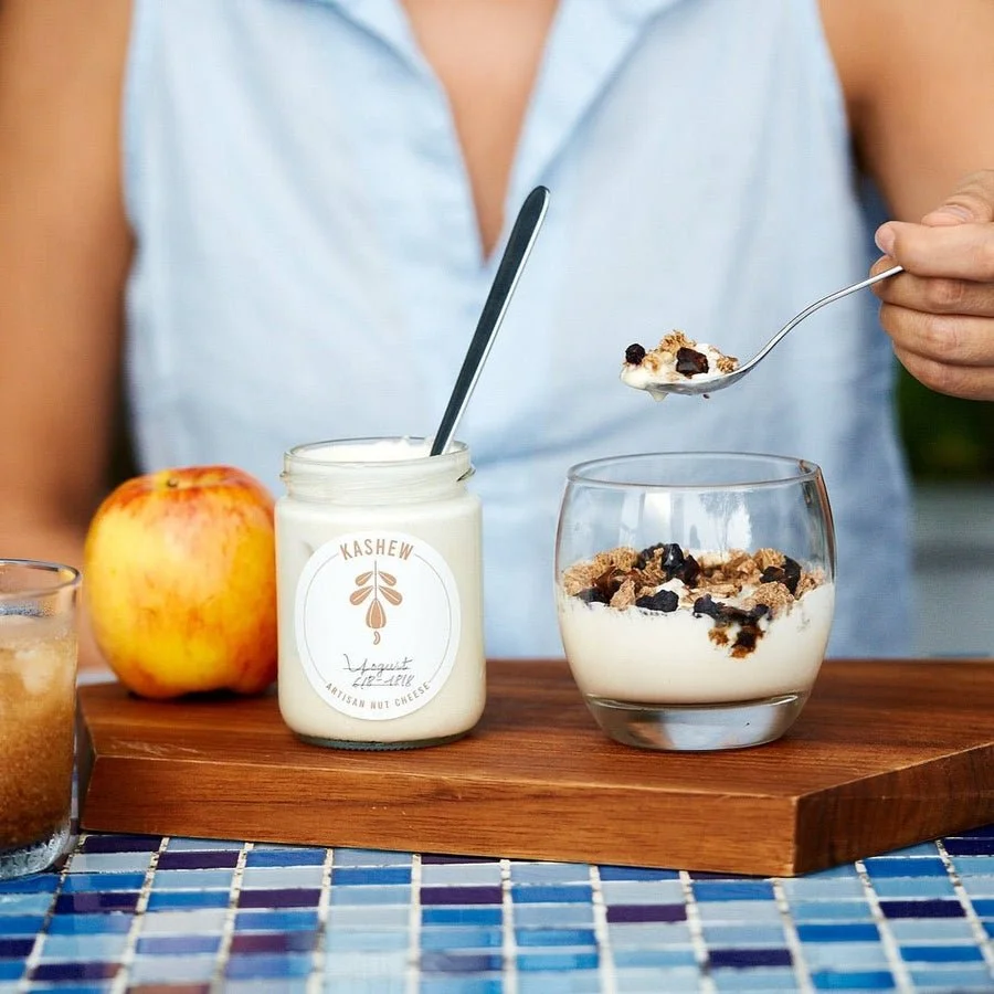 A person wearing a light blue sleeveless shirt is holding a spoonful of yogurt with granola and dried fruit over a glass bowl filled with yogurt and granola. There is a jar of yogurt labeled "Kashew Yogurt," a red apple, and a glass of iced beverage 