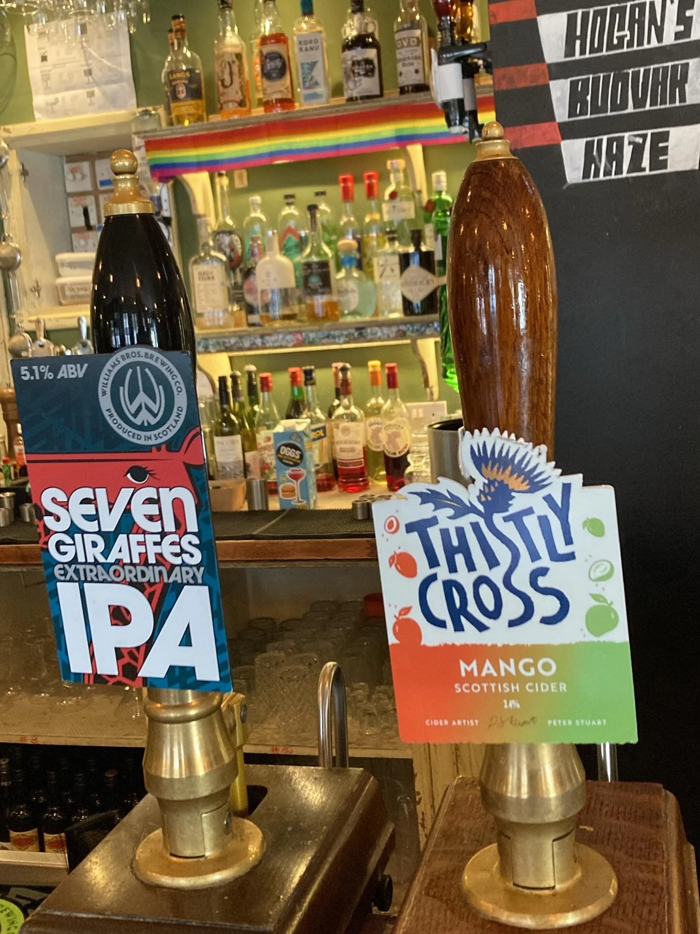 Bar scene with two beer tap handles, one black labeled 'Seven Giraffes Extraordinary IPA' and one wooden with a sign reading 'Thistly Cross Mango Scottish Cider 4%.' In the background, shelves with various liquor bottles and a chalkboard listing diff