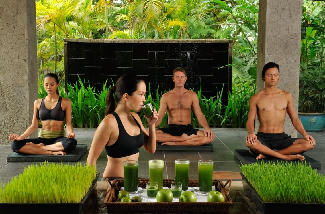 Four people practicing yoga outdoors surrounded by lush greenery, with a woman drinking a green juice in the foreground, and glasses of green juice and green apples on a tray in front of her.