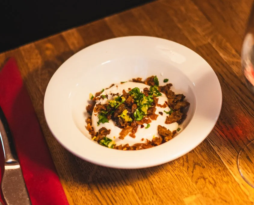 Bowl of creamy dip topped with chopped herbs and crumbled cooked meat, served on a wooden table.