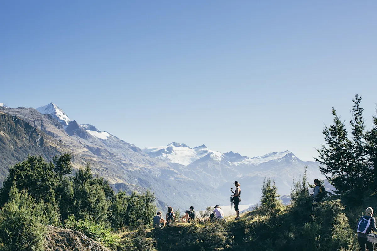 Group of hikers resting on a grassy hillside with snow-capped mountains in the background on a clear day.