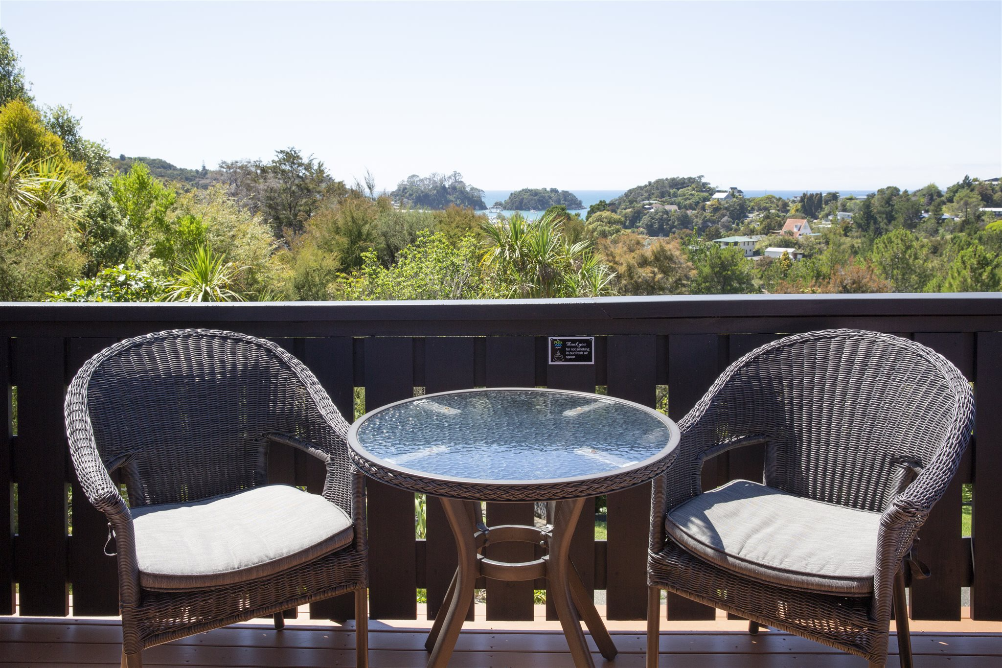 A balcony with a glass-top round table and three wicker chairs, overlooking a lush green landscape with trees, houses, and a distant body of water under a clear blue sky.