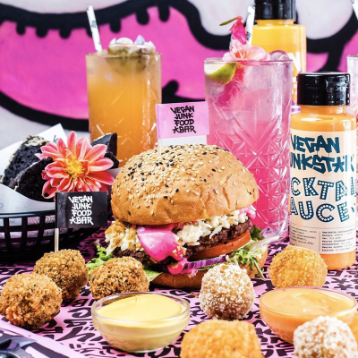 A vegan burger with fried chicken, lettuce, tomato, and pickles in a bun, surrounded by fried balls and dipping sauces. Two colorful drinks and a bottle of orange drink sit behind the burger, with flowers and a small sign that reads "Vegan Junk Food 