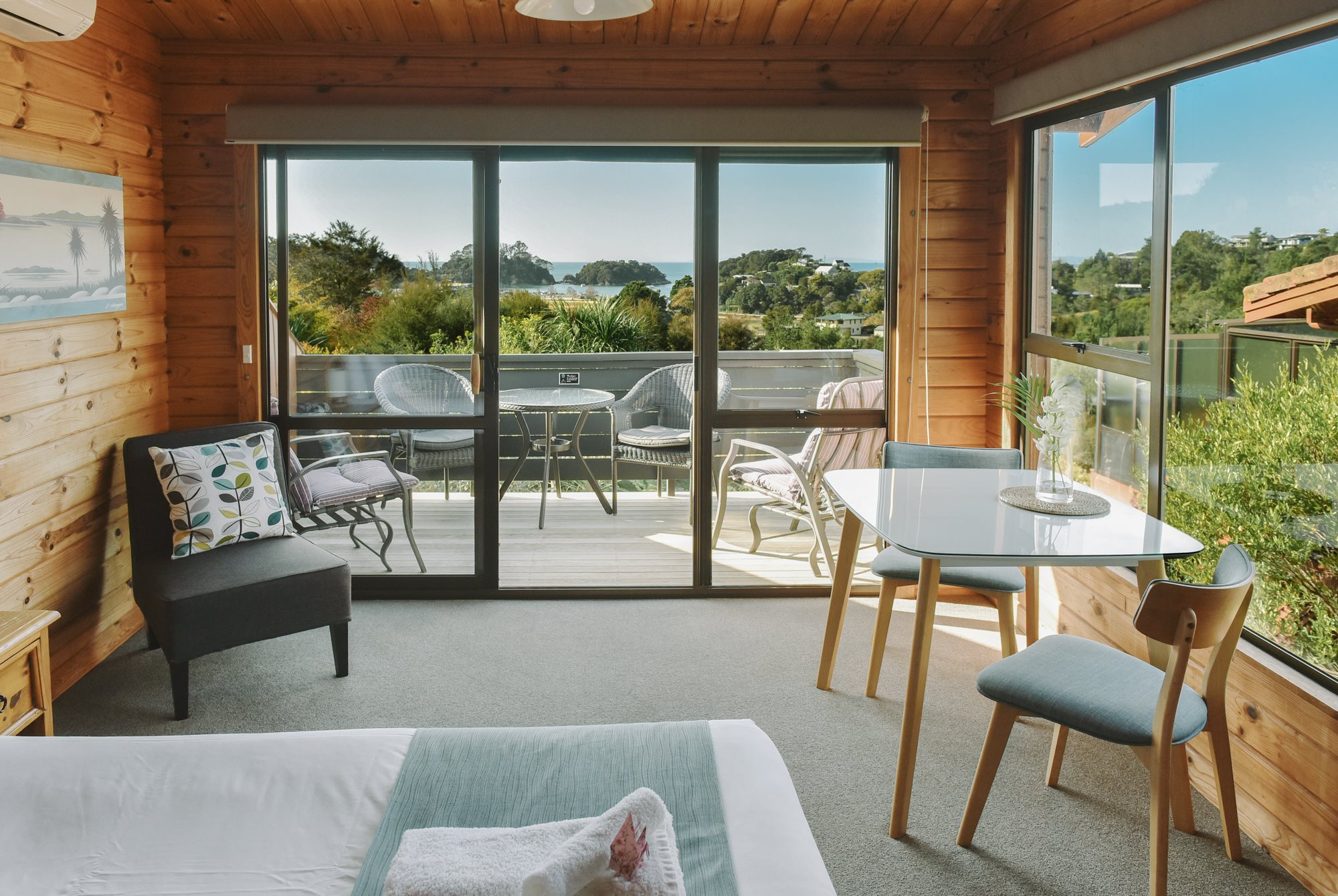 A cozy living room with wood-paneled walls and large sliding glass doors leading to a balcony with outdoor seating. The view shows lush greenery, a body of water, and distant hills under a clear blue sky.