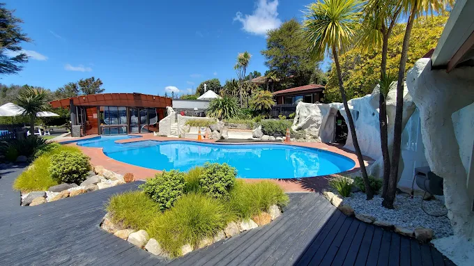 Outdoor swimming pool surrounded by lush landscaping, palm trees, and a clear blue sky with some white clouds.