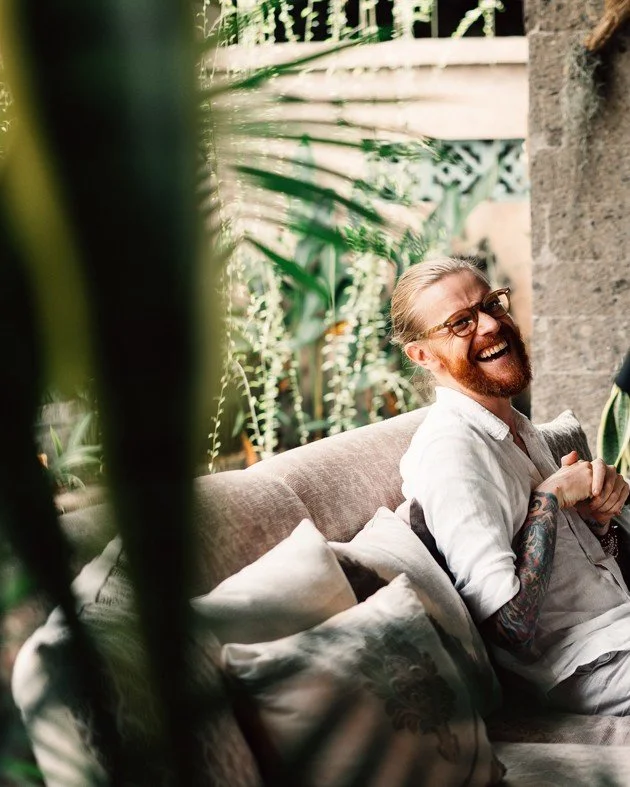 A man with glasses and tattoos sitting on a sofa, laughing and holding a cup, in a cozy indoor space with plants and stone wall in the background.