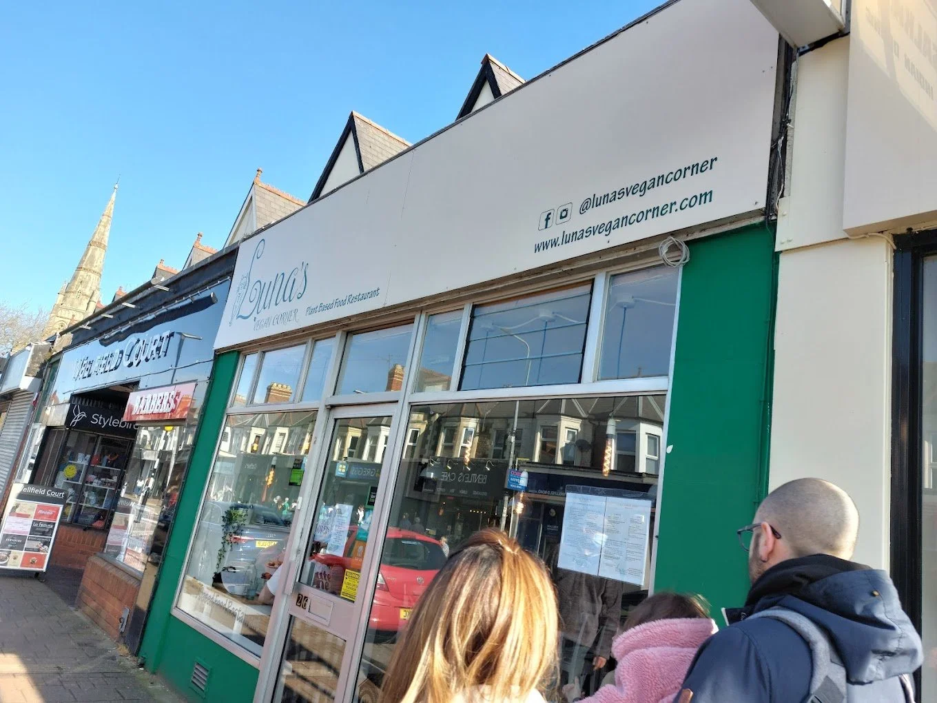 People waiting outside a restaurant on a city street under a clear blue sky.