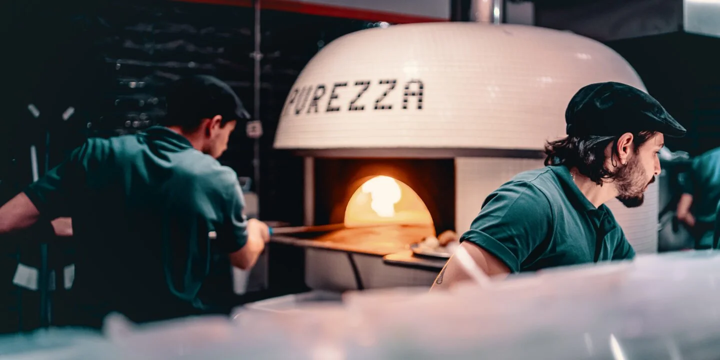 Two pizza chefs placing a pizza into a wood-fired oven with the word 'PUREZZA' on the oven's dome.
