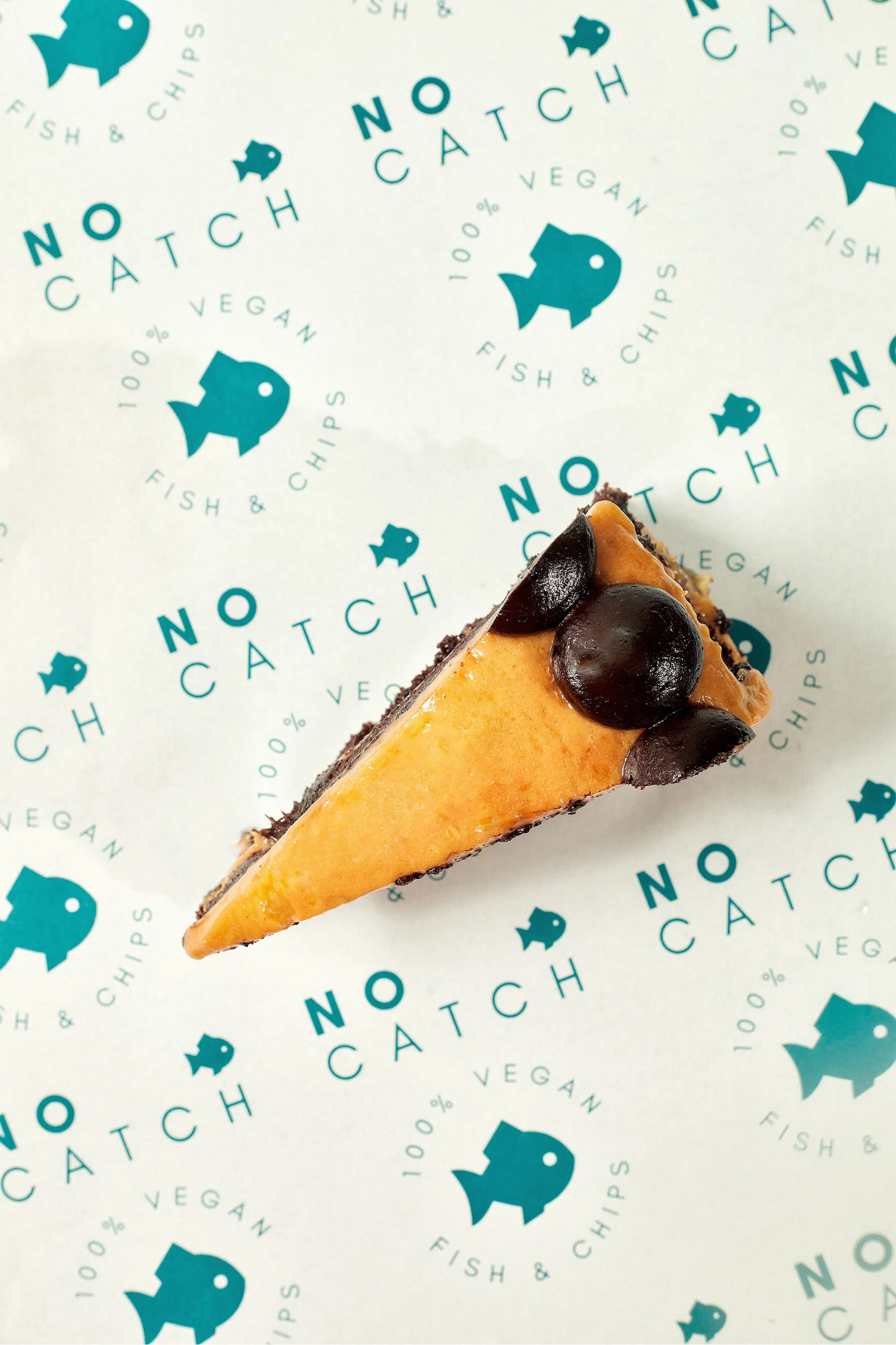 A slice of cheesecake with a golden top and chocolate chips on top, on patterned paper with blue fish and the words "NO CATCH" and "FISH & CHIPS".