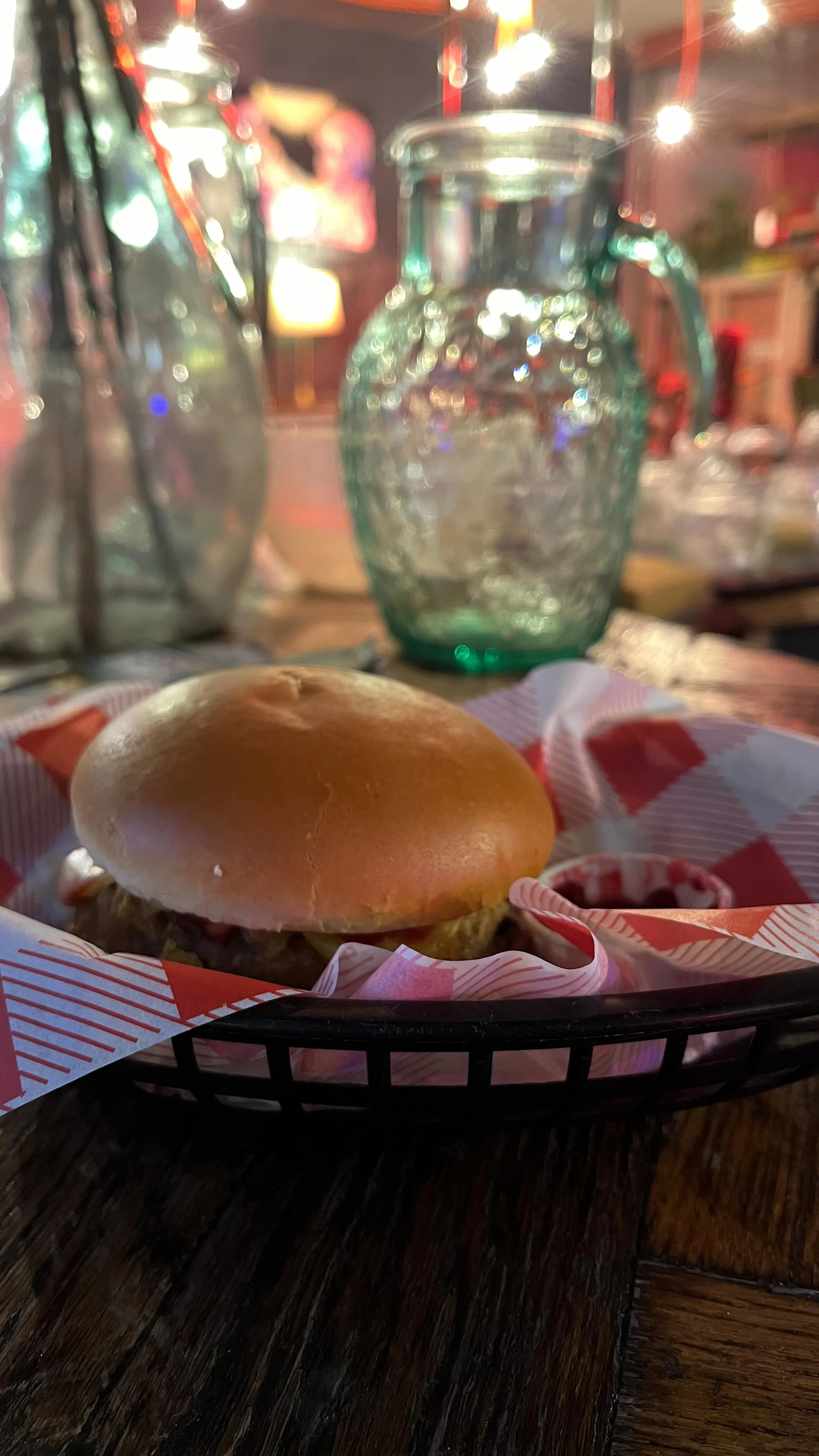 A burger in a basket with red and white checkered paper, a small container of ketchup, with a glass pitcher, a water bottle, and blurred background lights and decor.