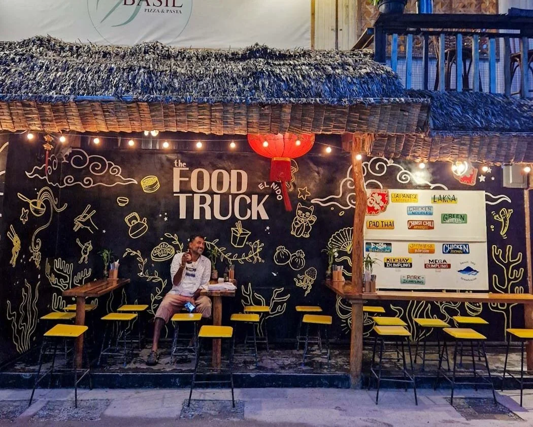 Outdoor food truck seating area with a man sitting at a table, holding a drink. The space is decorated with a black wall, yellow stools, and string lights. A menu board lists various Asian dishes.
