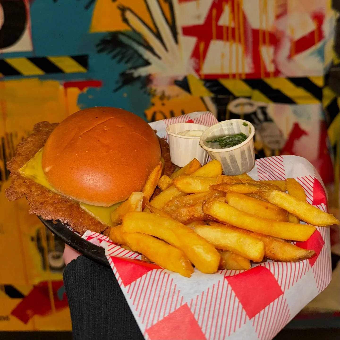 A serving of food including a cheeseburger with a bun, fried chicken cutlet, and melted cheese, French fries, and two small cups of condiments, served on a red and white checkered paper tray.