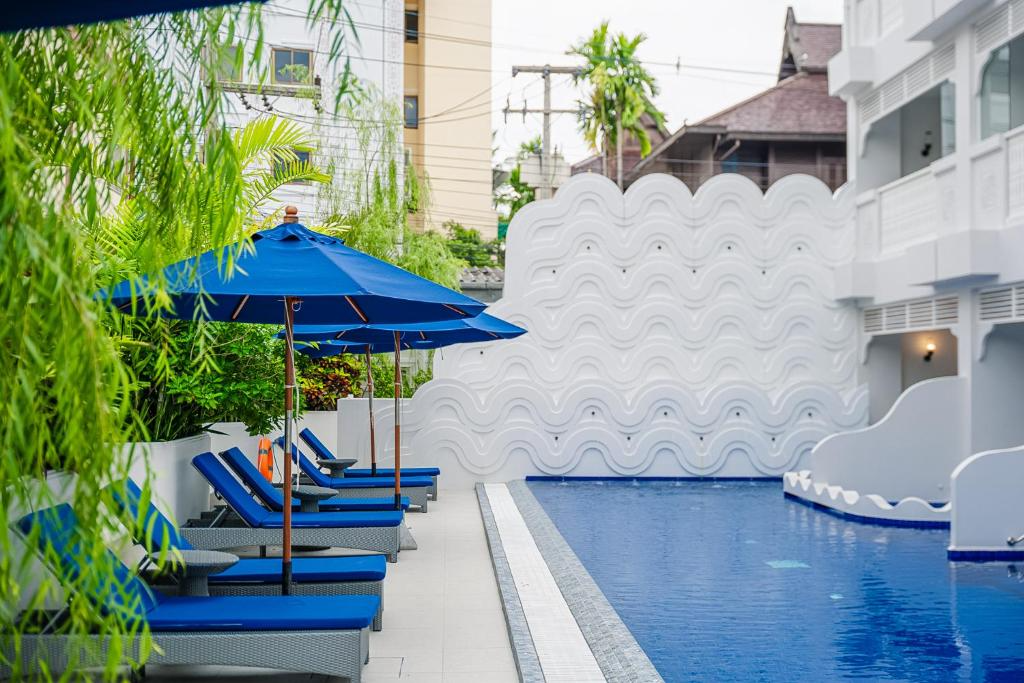 Empty outdoor swimming pool with blue lounge chairs and umbrellas, lush green plants, and a white decorative wall in the background.