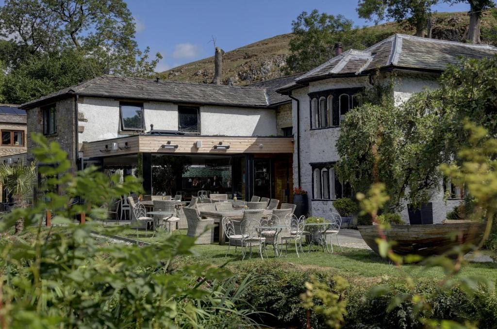 A large house with white stucco walls and black trim, featuring multiple windows and a patio with outdoor furniture, surrounded by greenery and a hillside in the background.