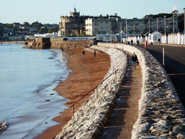 A coastal walkway with a stone wall, people walking along it, sandy beach with waves, historic castle-like building in the background, and a row of colorful beach huts or structures.