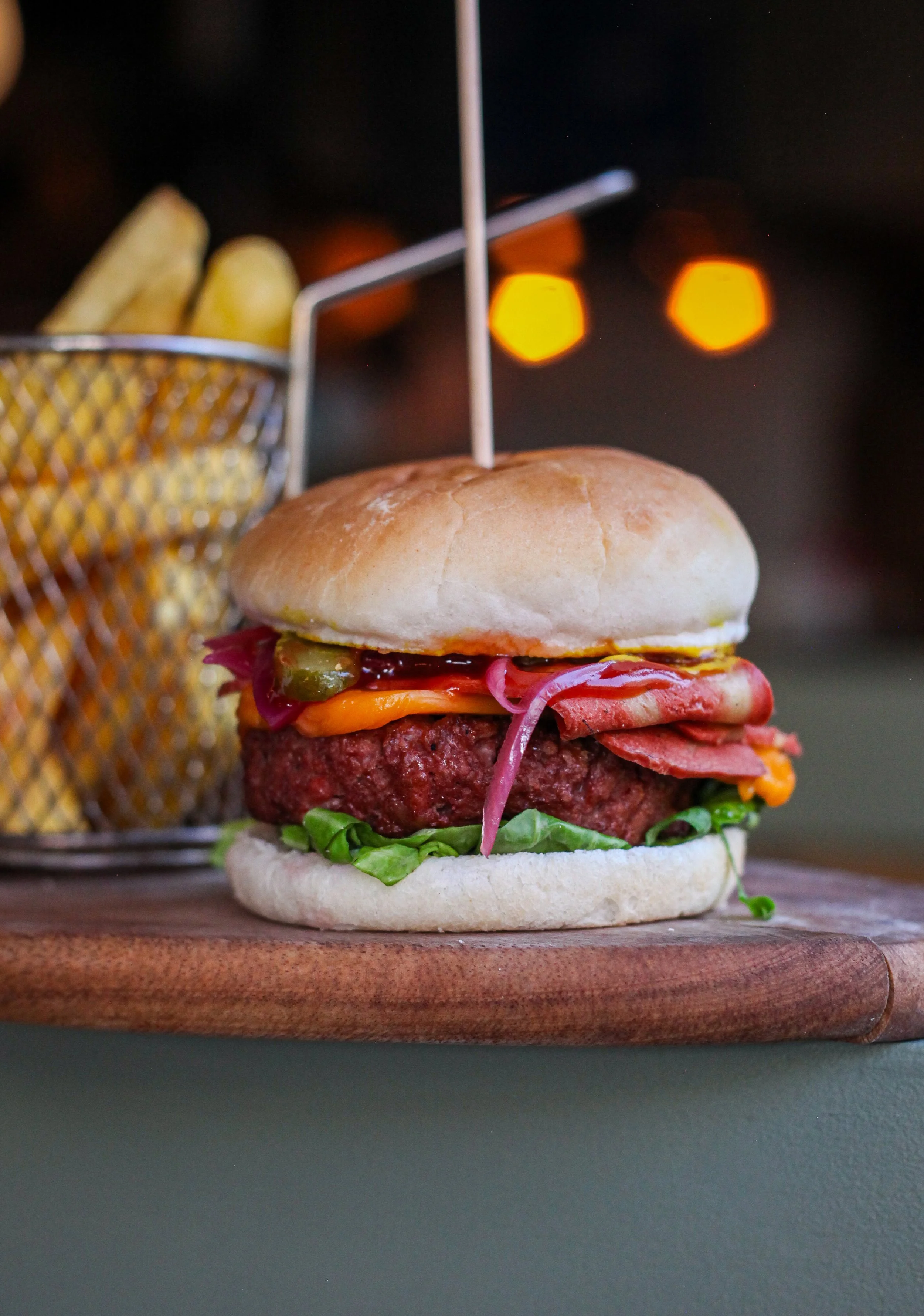 Close-up of a gourmet burger with lettuce, tomato, pickles, onion, and melted cheese in a bun, placed on a wooden cutting board. In the background, there are potato fries in a metal basket and warm bokeh lights.