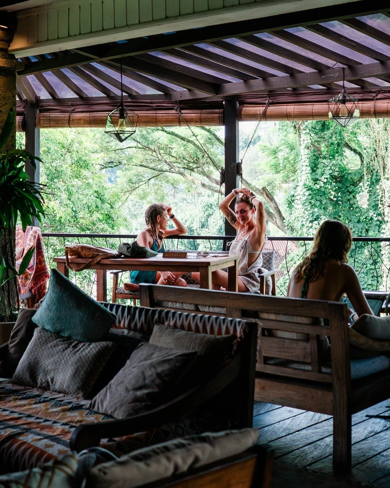 People sitting on a covered outdoor patio with lush greenery in the background. There are three women at a table, one is adjusting her hair and smiling at the camera, the other two are engaged in conversation after sitting down. The area is decorated