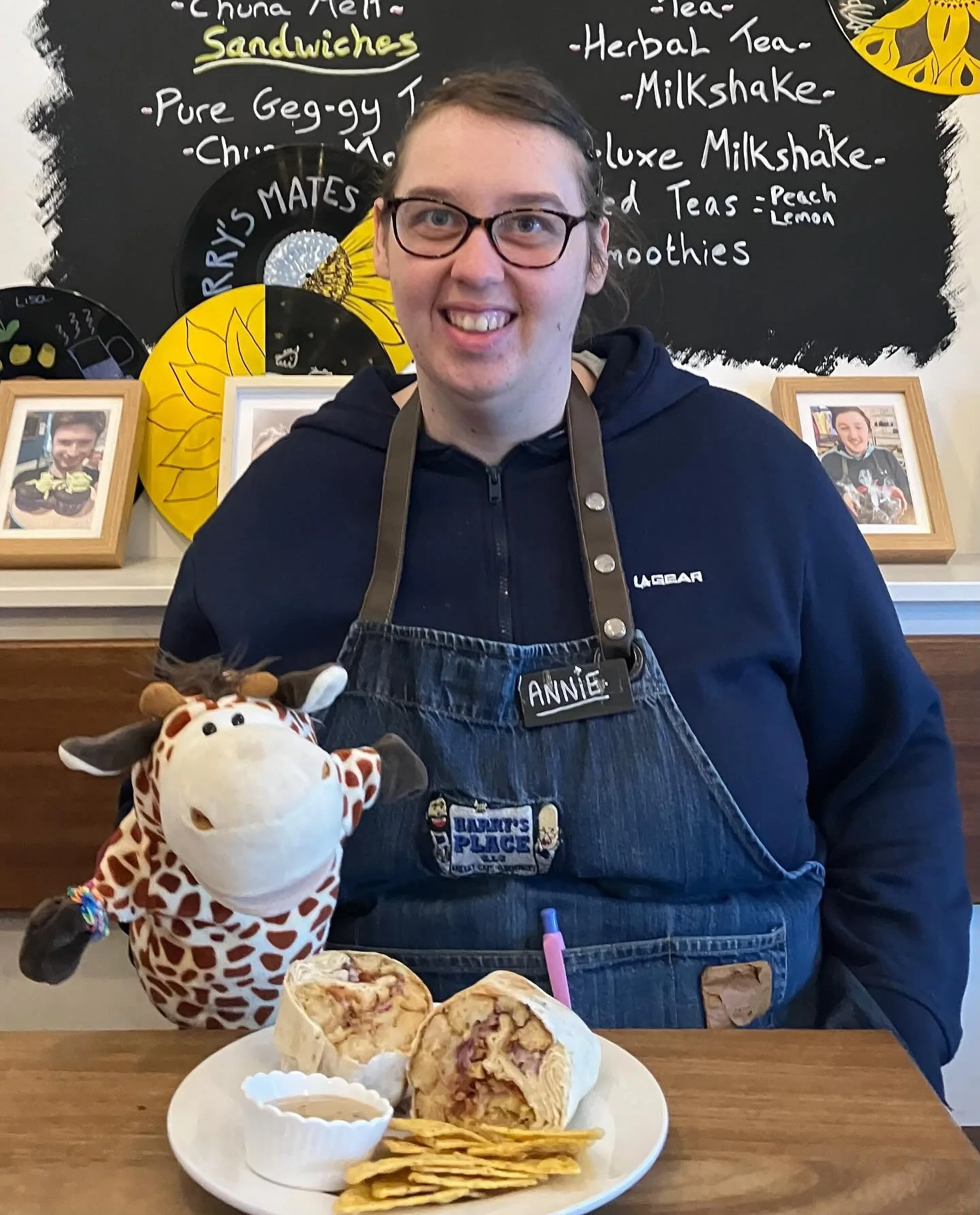 A woman with glasses and a name tag that says Annie, wearing a blue apron, is sitting at a table with a plate of food, a stuffed giraffe toy, and a burrito. She is smiling in front of a chalkboard menu listing sandwiches, teas, milkshakes, and smooth