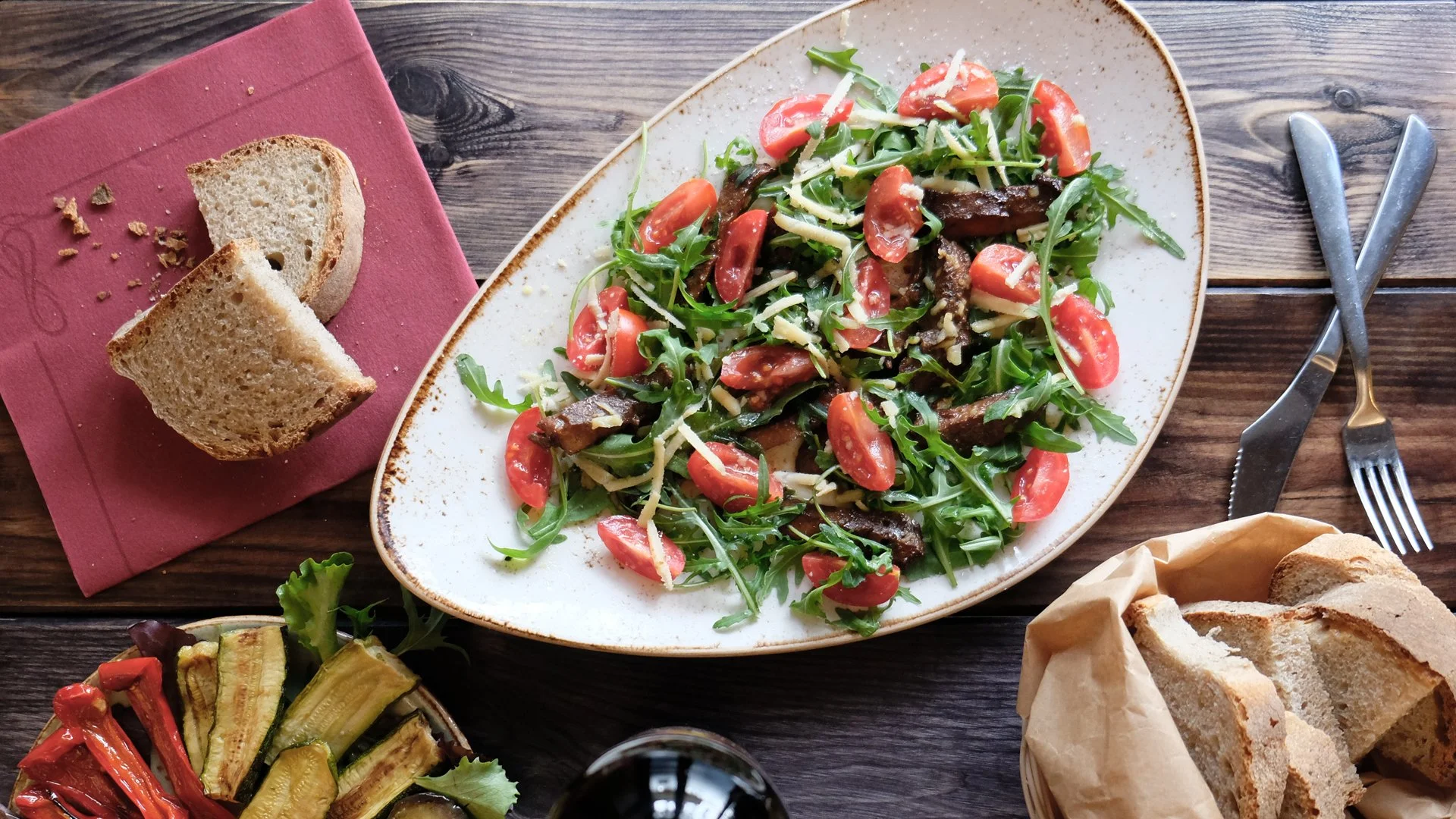 A plate of arugula salad with cherry tomatoes, grated cheese, and grilled meat slices on a rustic wooden table. Surrounding the plate are slices of bread, a fork and knife, a glass of red wine, and bowls of grilled vegetables.