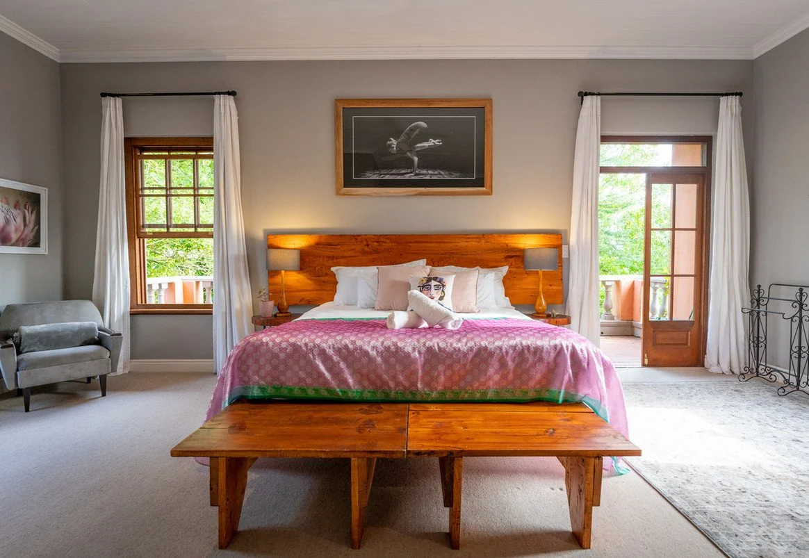 A bedroom with a large bed covered in a pink blanket, a plush toy, two pillows, and a wooden headboard. There are two lamps on matching bedside tables, a window and a door leading to a balcony, a gray armchair, a framed black and white dance photo above the bed, and a window with white curtains.