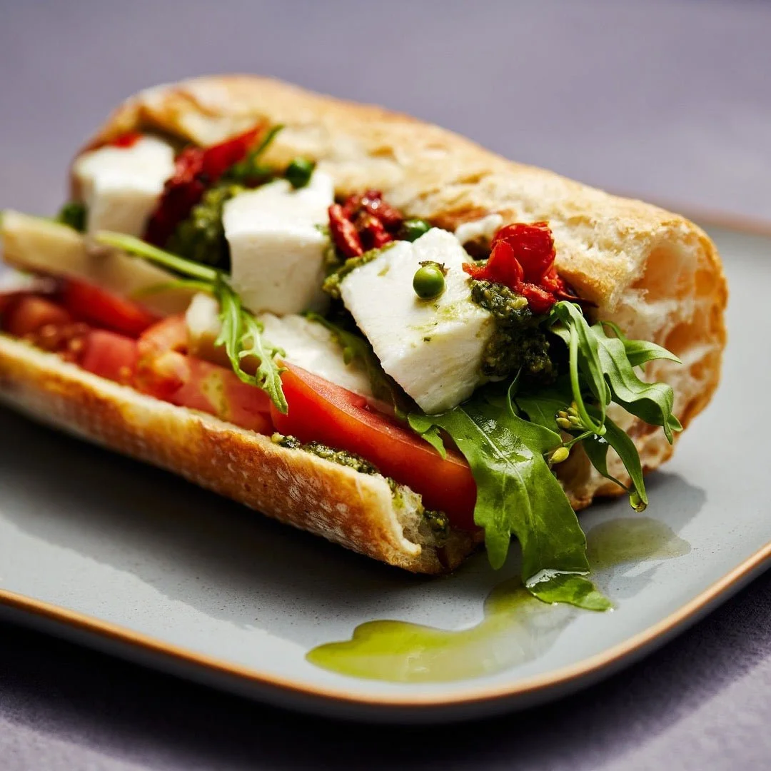 A close-up of a sandwich with fresh vegetables, feta cheese, and pesto sauce on a plate.