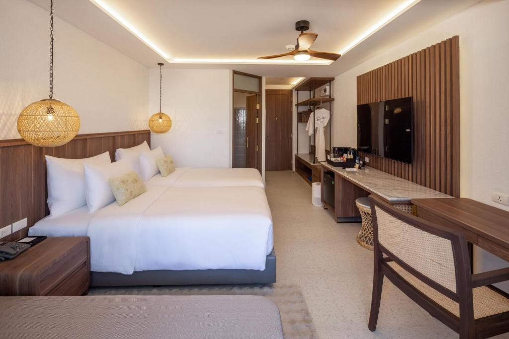 Hotel room with a large bed, wooden headboard, two hanging woven lamps, a flat-screen TV on a wooden panel, a desk with a chair, a closet with hangers, and a ceiling fan, decorated in neutral tones.
