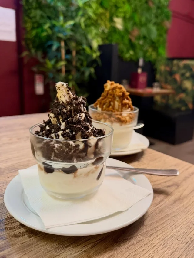 Two glasses of ice cream sundaes with toppings on a wooden table in a cafe.