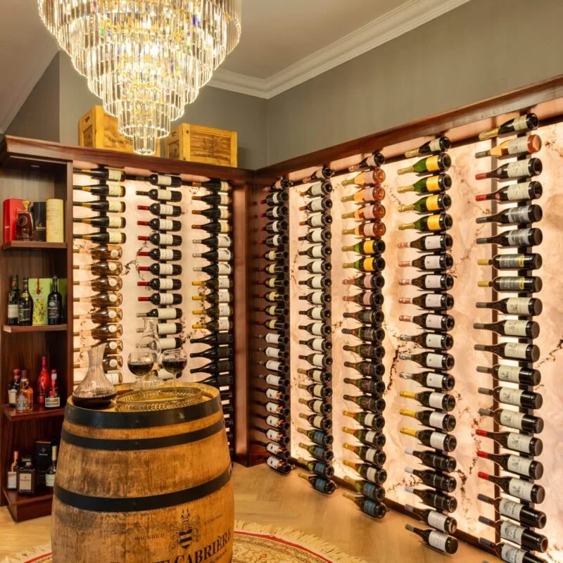 Wine bottles stored on wooden wine racks in a room, with a chandelier overhead, a barrel with wine glasses, and shelves of various wine bottles.