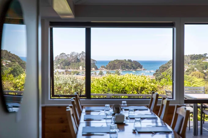 Dining table set for a meal inside a restaurant overlooking a scenic coastal view through large windows, with lush greenery and a bay with boats outside.