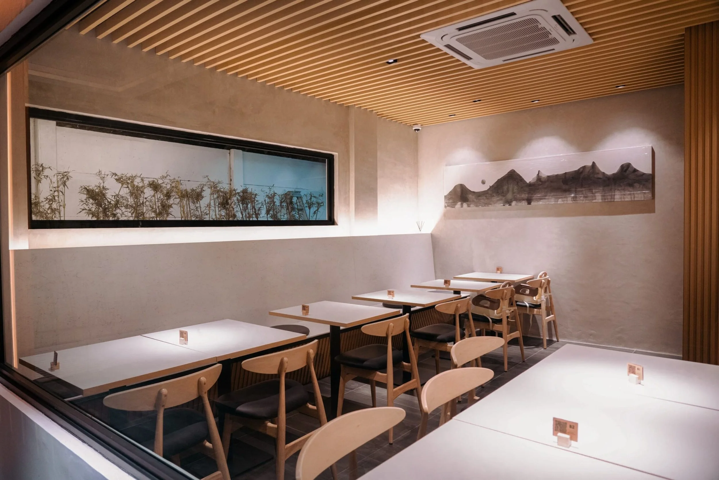 Interior of a modern restaurant or cafe with wooden ceiling slats, white tables, and wooden chairs, viewed through a large window.