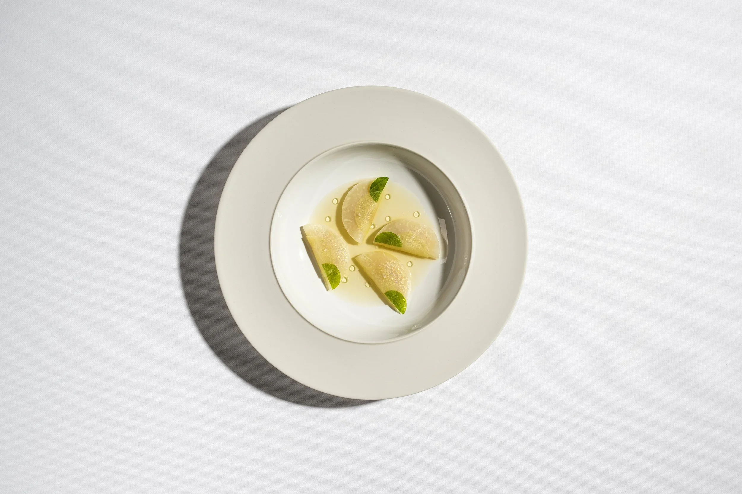 A white plate with lemon slices, small green leaves, and droplets of oil on a white surface.
