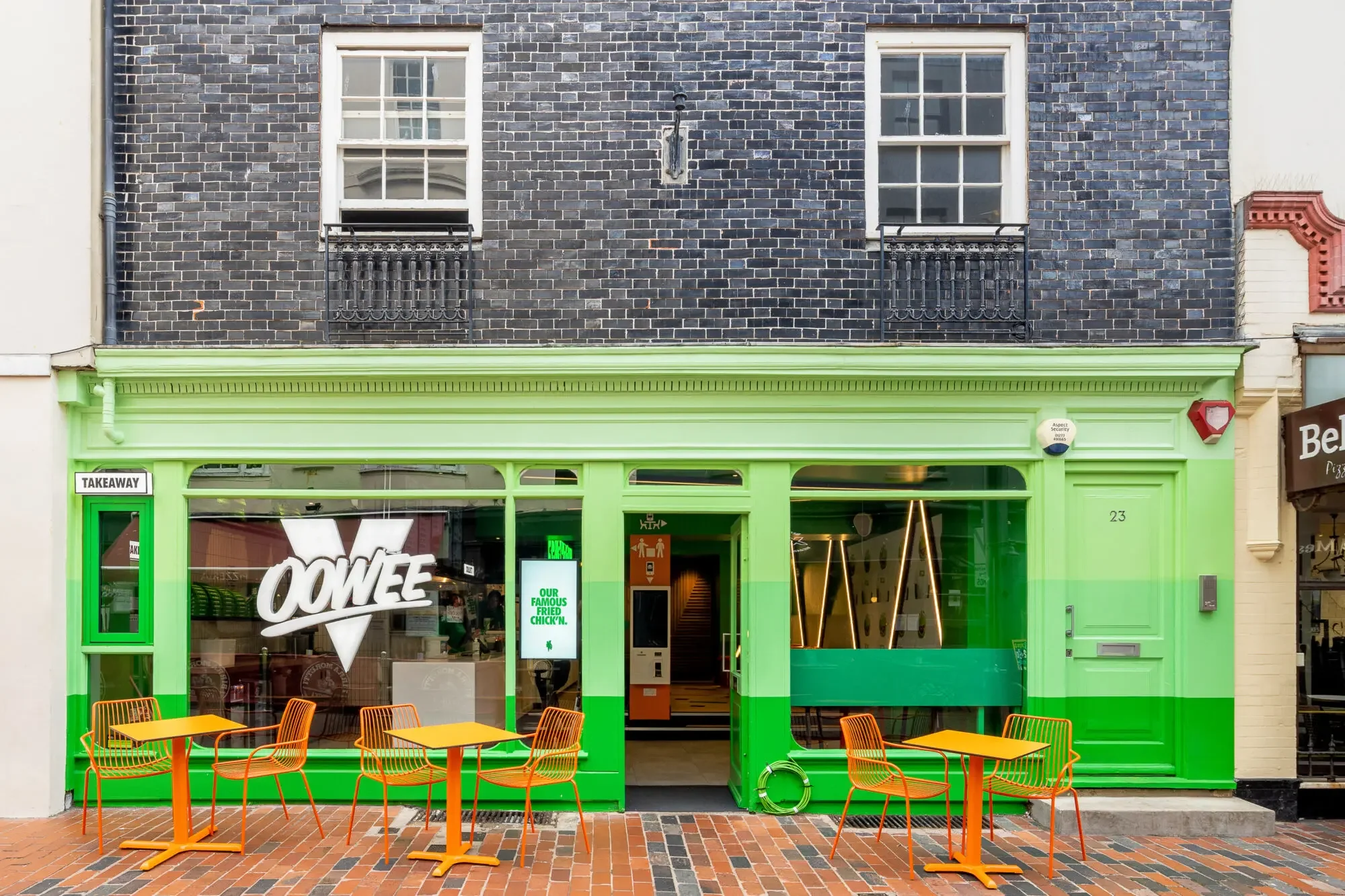 The storefront of a restaurant with bright green framing around the entrance. There are several orange tables and chairs outside on a brick sidewalk. The restaurant has a large window with the word 'OME' visible, a sign that reads 'OUR FAMOUS FRIED CHICKEN,' and a doorway leading inside.