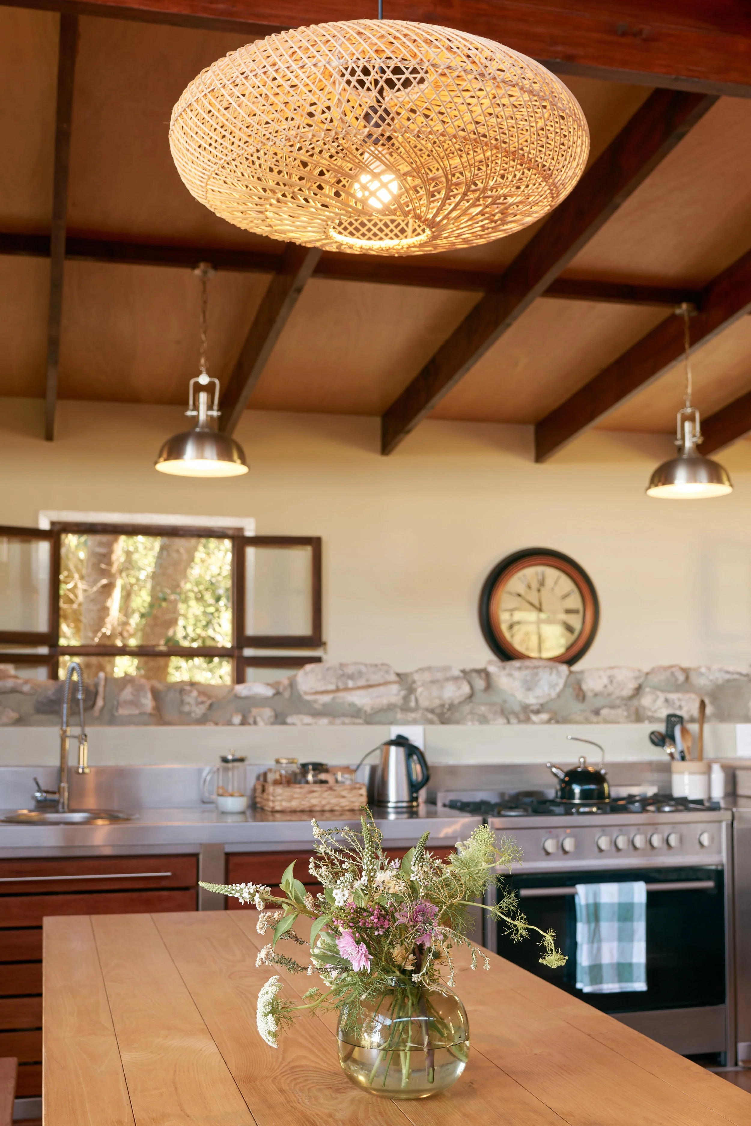 A kitchen with a wooden dining table, a vase with flowers, a stone backsplash, and a stainless steel stove. A woven rattan ceiling light hangs above, and two metallic pendant lights are on the ceiling. Large window with natural light and a clock on t
