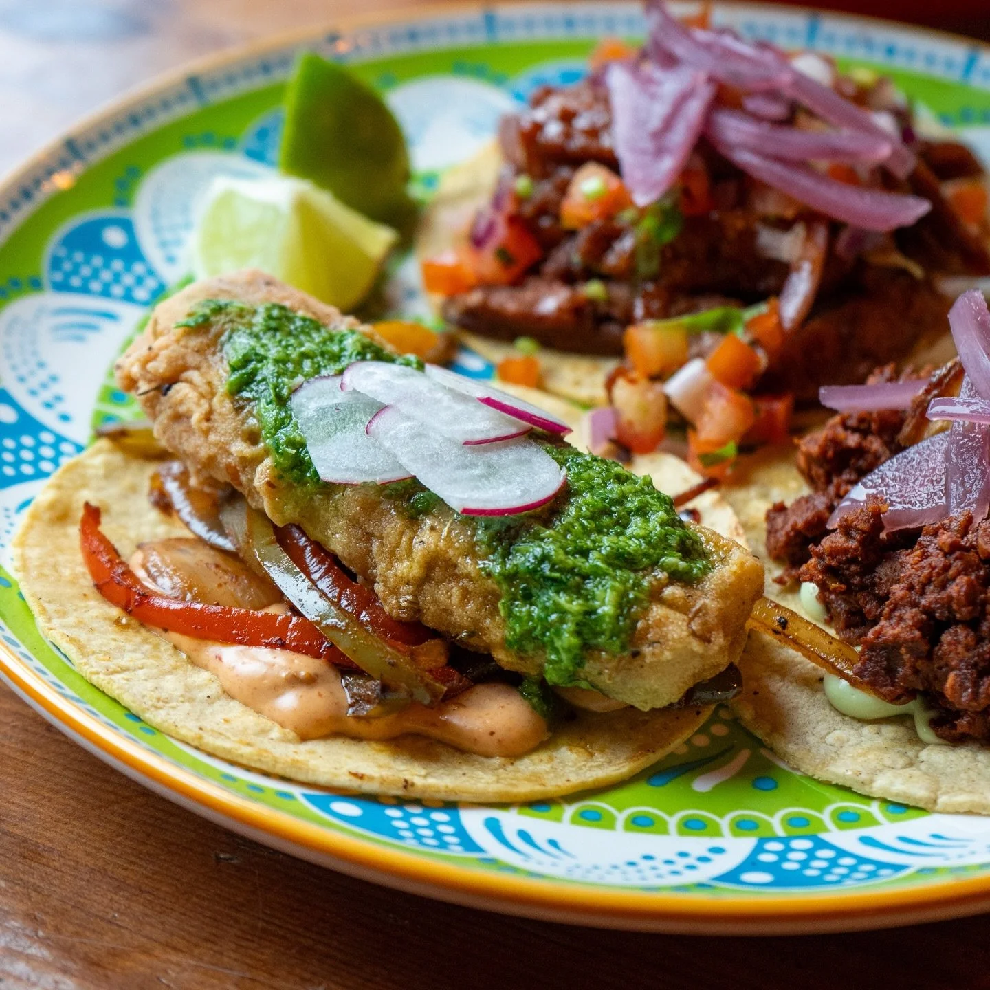 Close-up of two tacos with fried fish, sliced radishes, topped with green sauce, and placed on corn tortillas. In the background, there are beef tacos with chopped onions and cilantro, and beef tinga tacos with red onions on top, all on a colorful plate with lime wedges.
