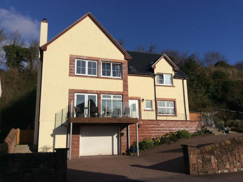 A multi-story house with cream-colored walls, red brick accents around the windows, and a black roof. There is a terrace with glass railing and outdoor furniture, and a garage door at the bottom. The house is set on a sloped yard with stone retaining walls and stairs, with a hillside covered in trees in the background.
