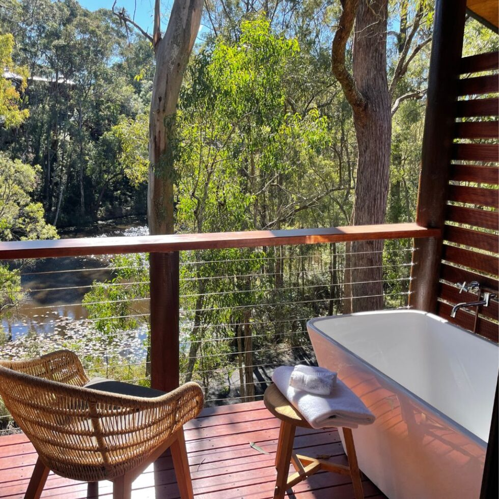 Outdoor bathtub on a balcony overlooking a wooded river area with sunlight filtering through trees, a wicker chair, and a small table with towels.