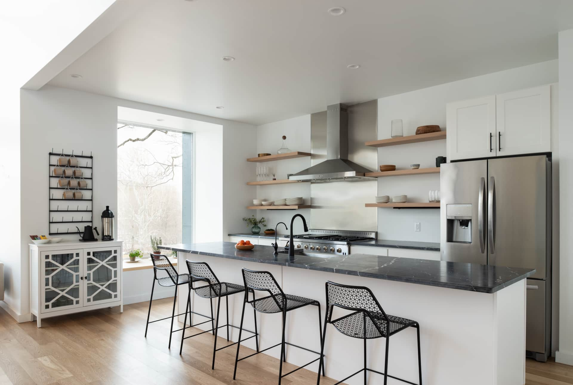 Modern kitchen with white walls, wooden open shelves, stainless steel appliances, black faucet, black barstools, and a large window.