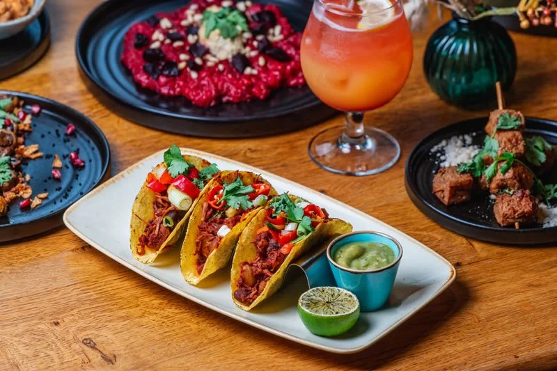 A white rectangular plate with three tacos filled with shredded meat, topped with chopped red and green peppers and cilantro, garnished with lime, alongside a small cup of green sauce. In the background, there are various other plates with colorful dishes and drinks on a wooden table.