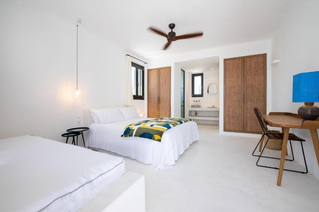 A bright, minimalistic bedroom with white walls and bedding, wooden furniture, a ceiling fan, and a small desk with a chair and a blue lamp.