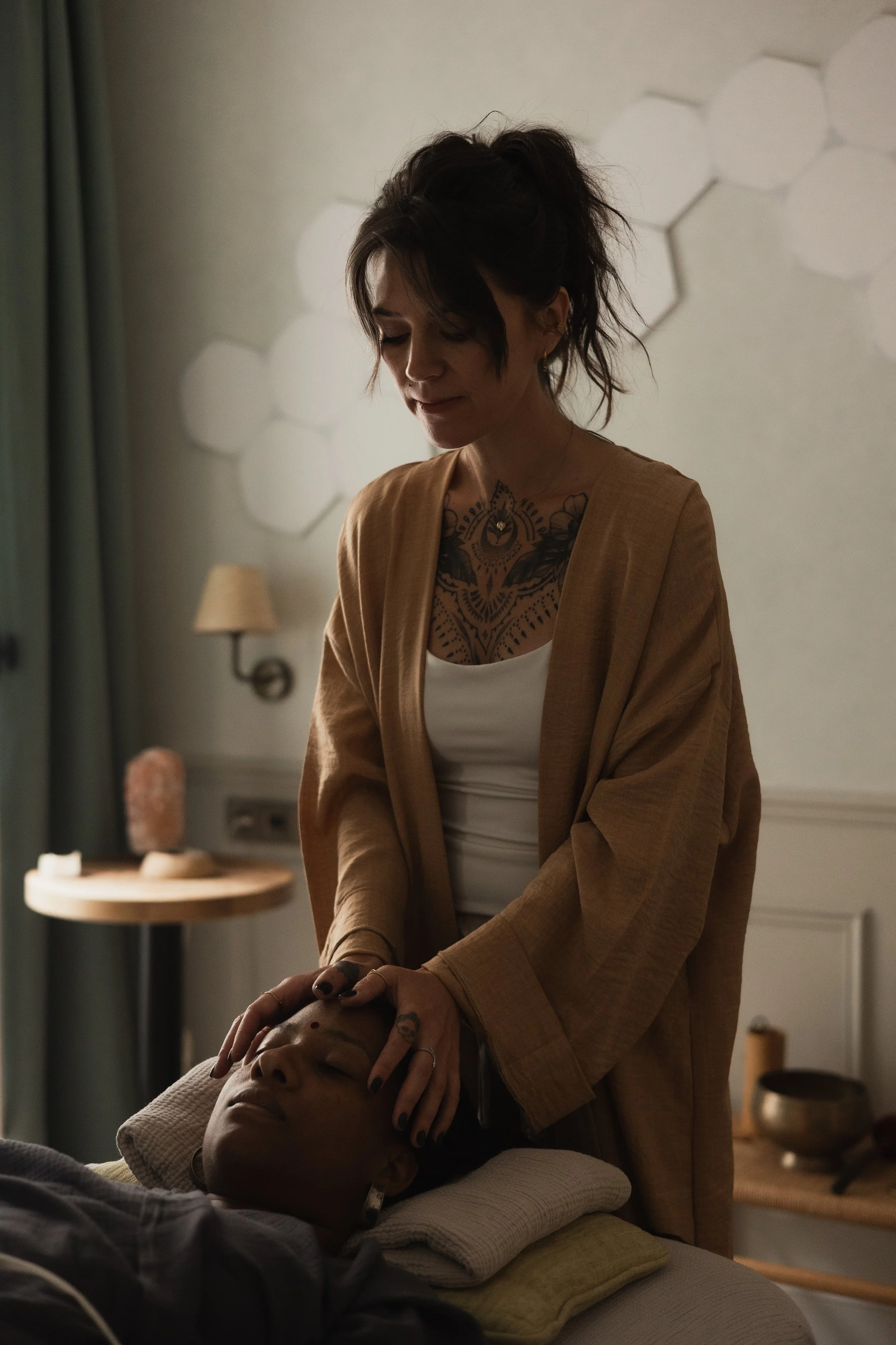 A woman giving a facial massage to a person lying on a massage bed in a softly lit room.