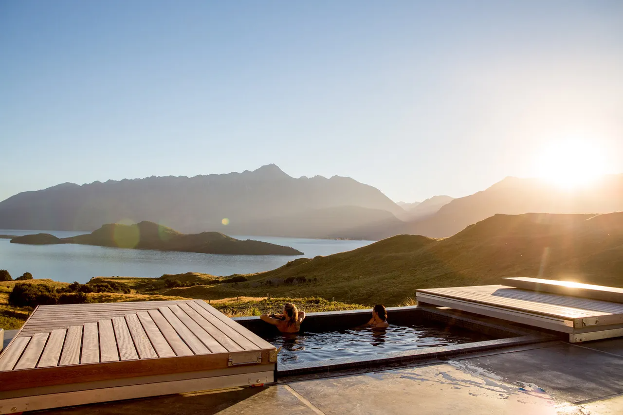 Two people relaxing in an outdoor hot tub overlooking a scenic lake and mountain landscape during sunset.